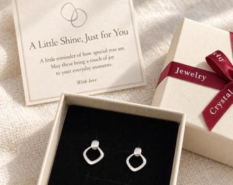 Könnte beinhalten: Ein Paar silberfarbene geometrische Ohrstecker in abgerundeter Rautenform, präsentiert in einer offenen beigen Geschenkbox mit schwarzem Samteinsatz. Eine quadratische Karte enthält den Text A Little Shine, Just for You.