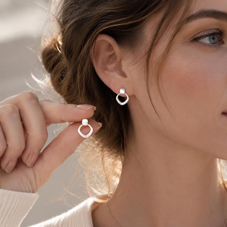 May include: A pair of silver-toned stud earrings featuring a small, smooth, rounded square top connected to a larger, open, teardrop-shaped bottom element. The earrings have a polished metallic finish and a simple, geometric design.