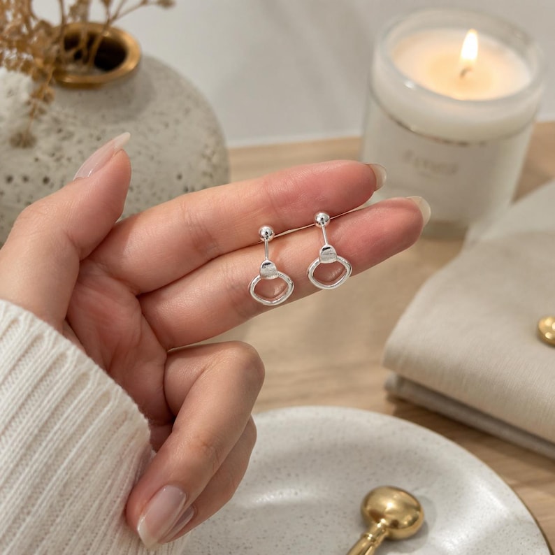 May include: A pair of silver stud earrings featuring a small polished sphere attached to a delicate, open circular drop design. The earrings have a bright, reflective metallic finish and a simple post-back mechanism for secure attachment to the earlobe.