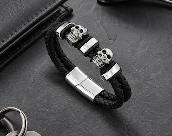 May include: A black braided leather bracelet featuring two silver-toned skull charms and three polished silver-toned metal spacers. The bracelet is secured with a rectangular silver-toned magnetic clasp, creating a layered look with its dual-strand design.