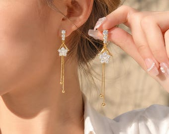 Könnte beinhalten: Goldfarbene Ohrhänger mit einem kleinen Stecker, einem sternförmigen Anhänger mit klaren Kristallen und zwei zarten, herabhängenden Goldketten, die in kleinen goldenen Kugeln enden, was ein geschichtetes und bewegliches Design erzeugt.