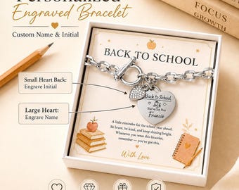 Könnte beinhalten: Ein silberfarbenes Gliederarmband mit Knebelverschluss und zwei Herzanhängern in einer weißen Geschenkbox. Ein Herz ist klein und texturiert, das andere ist groß und mit "Back to School", "You've Got This" und "Francis" graviert. Die Box enthält Text.