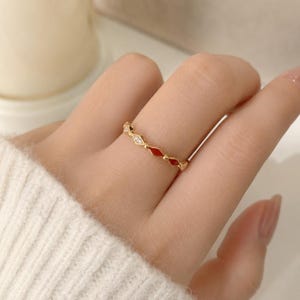Puede incluir: Un anillo fino de color dorado con un patrón repetitivo de pequeñas piedras claras y brillantes que se alternan con incrustaciones de esmalte rojo vibrante en forma de diamante, creando un diseño de banda delicado y simétrico en el dedo.
