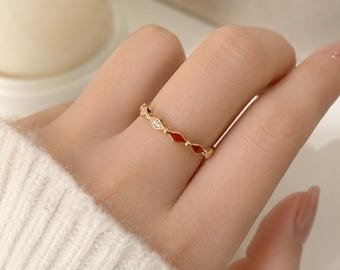 Könnte beinhalten: Ein dünner goldener Ring mit einem sich wiederholenden Muster aus kleinen, klaren, funkelnden Steinen, die sich mit glatten, leuchtend roten, rautenförmigen Emaille-Einlagen abwechseln und ein zartes, symmetrisches Banddesign am Finger bilden.