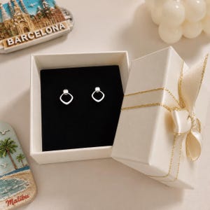 May include: A pair of silver-toned stud earrings featuring a geometric diamond-shaped outline. The earrings are displayed inside an open, cream-colored square gift box with a matching ribbon, set against a black velvet insert for contrast.