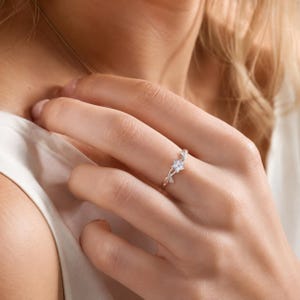 Puede incluir: Un anillo de tono plateado con un delicado diseño floral y una piedra clara central. La banda es fina y pulida, mostrando un pequeño motivo de flor con pétalos que se curvan en una estructura similar a una enredadera a lo largo del dedo.