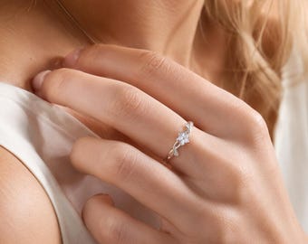 May include: A silver-toned ring featuring a delicate floral design with a central clear stone. The band is thin and polished, showcasing a small flower motif with petals that curve into a vine-like structure along the finger for a refined look.