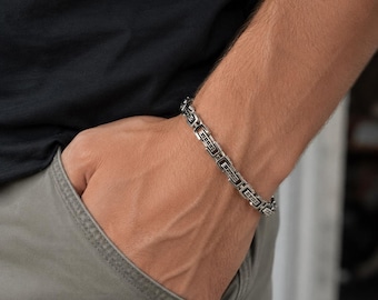 May include: A silver-toned metal chain bracelet featuring a series of rectangular links with a dark, recessed pattern. The bracelet has a polished finish and a secure clasp mechanism, worn around the wrist over a black shirt and gray pants.