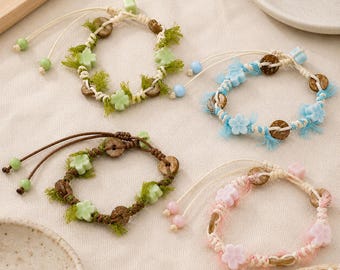 Könnte beinhalten: Vier verstellbare Armbänder mit geflochtenen Kordelbändern, runden braunen Holzperlen und pastellfarbenen blumenförmigen Anhängern. Jedes Armband enthält ausgefranste Stoffakzente in passenden Farben sowie zwei kleine Perlen an den Zugbändern.