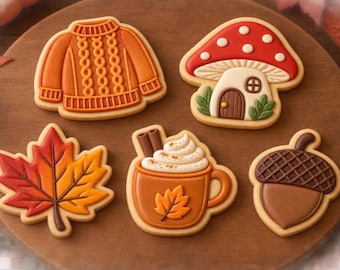 May include: Five decorated sugar biscuits shaped like a cable-knit jumper, a mushroom house, a maple leaf, a mug of hot cocoa with cinnamon sticks, and an acorn. The biscuits feature orange, red, brown, and white icing details.