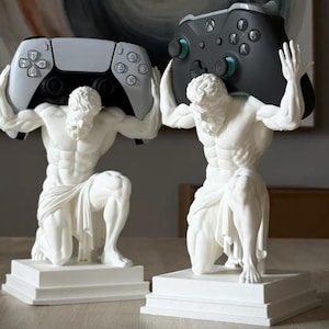 Puede incluir: Dos estatuas blancas de figuras arrodilladas con complexión musculosa, cada una sobre una base cuadrada. Una figura sostiene un mando de juego blanco sobre sus hombros, mientras que la otra sostiene un mando de juego negro.