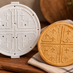 Op de afbeelding: Een ronde witte plastic vorm met een verhoogd kruisontwerp met kleine kruisaccenten in elk kwadrant, geplaatst naast een rond, goudbruin gebakken koekje dat hetzelfde reliëf kruispatroon op het oppervlak vertoont.