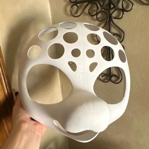Könnte beinhalten: Eine weiße, 3D-gedruckte Tiermaskenbasis mit einer abgerundeten Kopfform, einer ausgeprägten Schnauze, großen Augenöffnungen und einer Reihe kreisförmiger Ausschnitte im oberen Bereich, die eine sichtbare, geschichtete Textur aufweist.