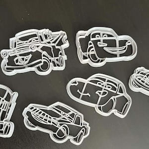 May include: A set of six white plastic cookie cutters shaped like various cartoon vehicles with expressive faces, including a tow truck, a race car, and other automotive designs, each featuring detailed outlines for cutting dough shapes.