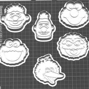 Op de afbeelding: Een set van vijf witte plastic koekjesvormen in de vorm van de gezichten van bekende kindertelevisiefiguren, waaronder Ernie, Bert, Elmo, Koekiemonster en Pino, gerangschikt op een donkergrijs oppervlak met een raster.