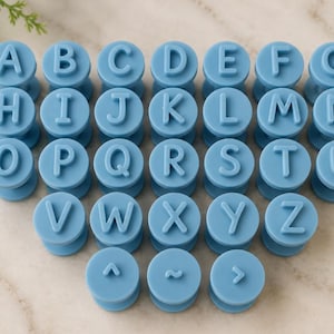 Puede incluir: Una colección de sellos circulares de color azul claro, cada uno con una letra mayúscula en relieve del alfabeto de la A a la Z, además de tres sellos adicionales con un acento circunflejo, una tilde y un símbolo de mayor que.