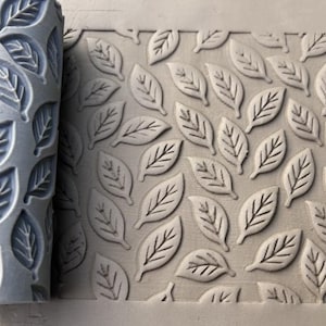 Puede incluir: Un rodillo de textura para arcilla gris de forma cilíndrica con un patrón de hojas repetitivo, colocado junto a una placa de arcilla gris que ha sido impresa con el mismo diseño de hojas, creando un motivo botánico en relieve uniforme.