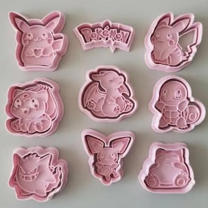 Op de afbeelding: Een set van acht lichtroze plastic koekjesvormen met diverse Pokémon-personages en een Pokémon-logo. De collectie bevat vormen van Pikachu, Bulbasaur, Charmander, Squirtle, Gengar, Pichu en Snorlax, elk met gedetailleerde contouren.
