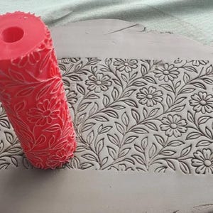Pode incluir: Um rolo de textura para argila de cor vermelho brilhante, de formato cilíndrico, com um padrão floral em relevo de margaridas e folhas. O rolo está posicionado sobre uma placa de argila cinzenta, exibindo um design estampado de flores.