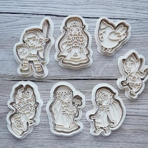 Peut inclure: Un ensemble de sept emporte-pièces en plastique blanc cassé représentant diverses formes de personnages fantastiques, dont un héros avec une épée, une princesse, un oiseau, une petite créature et d'autres figures stylisées pour la pâtisserie.