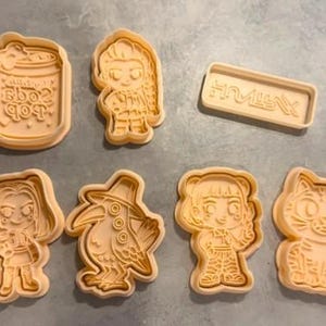 Pode incluir: Um conjunto de sete cortadores de biscoitos de plástico bege claro com vários designs, incluindo uma lata de refrigerante com o texto soda pop, três figuras humanas estilizadas, uma placa retangular com texto, um pássaro e um gato detalhado.