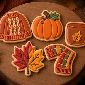 May include: Five decorated sugar cookies shaped like a cable-knit sweater, a pumpkin with a green leaf, a mug with a leaf design, a maple leaf with red and yellow icing, and a plaid scarf, all arranged on a round wooden serving board.