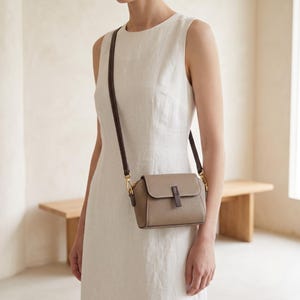May include: A small taupe crossbody bag with a textured finish, featuring a flap closure with a dark brown vertical tab detail, gold-tone hardware, and a thin dark brown adjustable shoulder strap, worn over a sleeveless white dress.