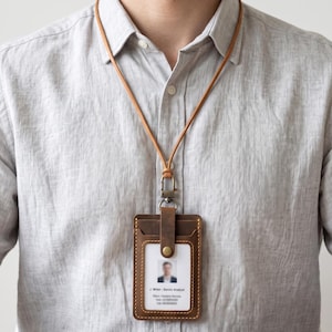 May include: A brown leather ID badge holder with a matching neck lanyard. The holder features a vertical orientation, visible stitching, a brass-colored snap button, and a metal swivel clasp. An identification card with a photo and text is inserted.