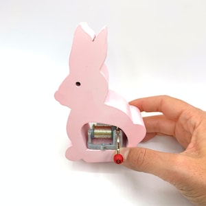 May include: A pink wooden bunny shaped music box with a wind-up key. The music box is made of wood and has a painted pink finish.