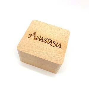 May include: A light-colored wooden box with the name "Anastasia" engraved on the lid. The box is square-shaped and has a natural wood grain finish, set against a white background.