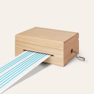 Può includere: Un carillon in legno chiaro con corpo rettangolare e una manovella in metallo argentato sul lato. Una lunga striscia di carta bianca con sottili linee verticali blu e un motivo a griglia fuoriesce da una fessura orizzontale sulla parte anteriore.