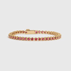 Puede incluir: Una pulsera de tenis en tono dorado que presenta una hilera continua de piedras redondas de color rosa vibrante, engastadas en biseles metálicos circulares individuales. La pulsera incluye un cierre de caja rectangular con lengüeta.