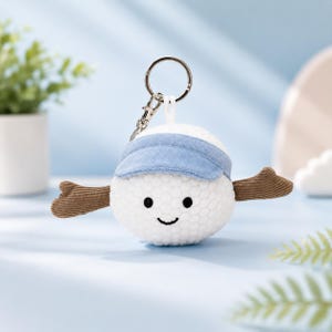 May include: A plush white golf ball keychain featuring a textured surface, a light blue fabric visor, and two brown corduroy arms extending from the sides. The keychain includes a silver metal ring and a lobster clasp for secure attachment.