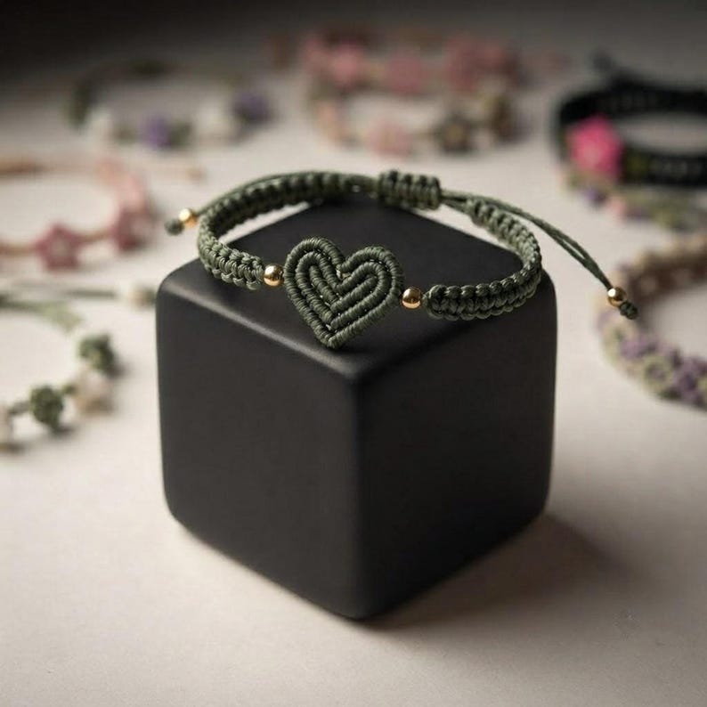 An olive green woven cord bracelet featuring a central heart motif and small gold-colored beads, draped over a black cube.