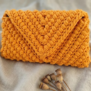 May include: A rectangular mustard yellow clutch bag crafted from thick, chunky crocheted fabric. The exterior features a prominent V-shaped chevron pattern created by the dense, interlocking stitches of the yarn, forming a textured surface.