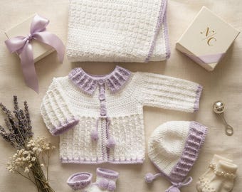 Puede incluir: Un conjunto de bebé de ganchillo en blanco y lavanda que incluye una manta, una chaqueta de manga larga con botones y pompones, un gorro y patucos. Se acompaña de una caja de regalo con lazo, un sonajero plateado y calcetines con perlas.