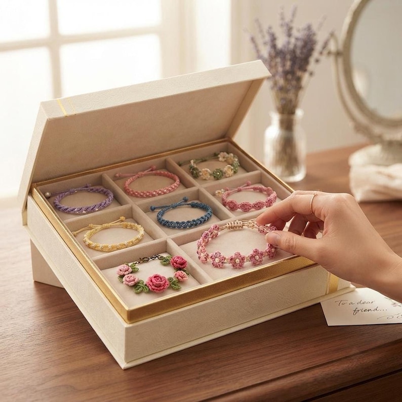 A hand reaching into an open beige jewelry box to touch a pink floral crochet bracelet among a collection of various colored bracelets.