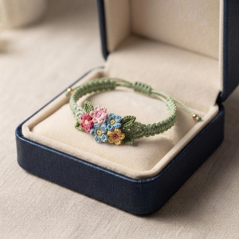 A light green crochet bracelet adorned with tiny pink, blue, and yellow flowers resting inside an open navy blue jewelry box.