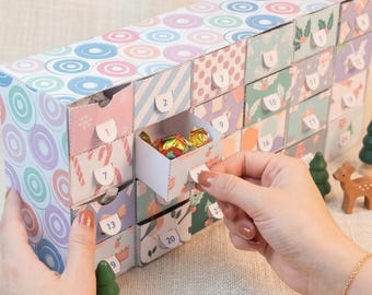 Puede incluir: Un calendario de adviento rectangular de cartón con veinticuatro cajones pequeños, cada uno etiquetado con un número. El exterior presenta un patrón colorido de círculos, rayas y motivos festivos. Un cajón está entreabierto, revelando un caramelo.