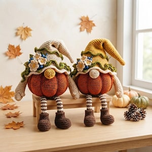May include: Two crocheted pumpkin gnomes with dangling legs, wearing floppy hats adorned with small white flowers and blue berries. The gnomes feature orange pumpkin bodies, striped legs, and brown boots, sitting on a small wooden bench.