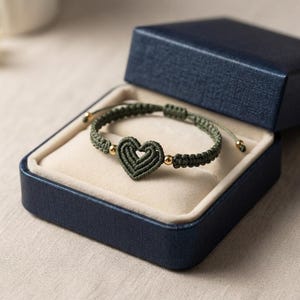 An olive green woven cord bracelet with a central heart charm and gold beads, resting inside an open blue jewelry box.