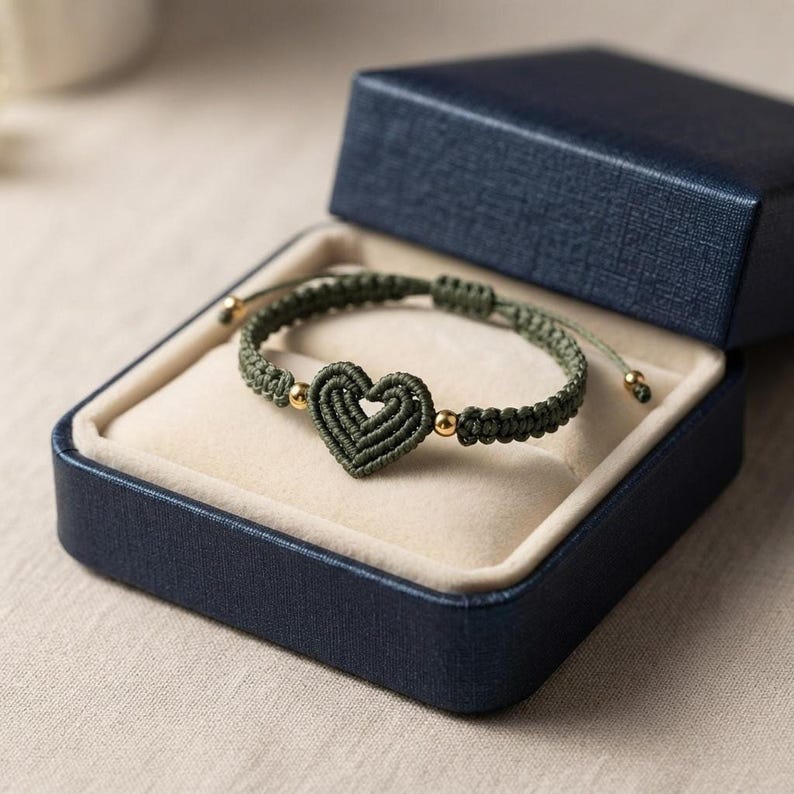 An olive green woven cord bracelet with a central heart charm and gold beads, resting inside an open blue jewelry box.