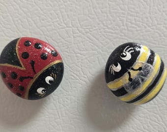 May include: Two painted rocks, one with a red and black ladybug design and the other with a black and yellow bee design.