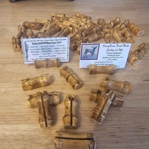 May include: A collection of natural bamboo place card holders. The holders are made of unfinished bamboo and are perfect for adding a rustic touch to any event. The image also includes two business cards with contact information for "DecorateWithBamboo.com" and "Honey Beach Hand Made Jewelry and Gifts".