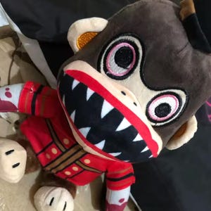 May include: A plush monkey toy featuring brown fur, a wide mouth with sharp white teeth, and large pink-rimmed eyes. The monkey wears a red jacket with gold accents and a black hat. A metal keyring is attached to the bottom of the plush toy.