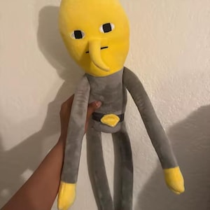 May include: A plush toy character with a tall, yellow, lemon-shaped head, a long nose, and small black eyes. The body is grey with long arms, yellow hands, a black belt with a yellow diamond-shaped buckle, and black feet at the end of long legs.