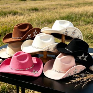 Pode incluir: Uma coleção de seis chapéus de cowboy em castanho, branco, preto, rosa e rosa claro. Cada chapéu apresenta uma fita a condizer e ornamentos prateados em forma de estrela ao longo da aba. Os chapéus estão dispostos numa superfície preta.