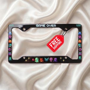 May include: A black rectangular license plate frame featuring pixelated video game monster graphics along the sides and bottom. The top center displays the text GAME OVER in a retro font, while the corners show SCORE +1000 in small white lettering.