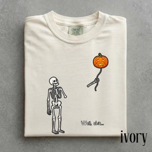 May include: An ivory t-shirt featuring a black skeleton graphic looking up at a floating orange jack-o'-lantern pumpkin held by a detached skeletal arm. The text "Well, shit..." is printed in black near the bottom right of the graphic design.