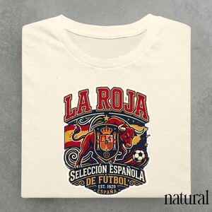 Può includere: Una t-shirt color naturale con una stampa grafica che riporta il testo LA ROJA SELECCIÓN ESPAÑOLA DE FÚTBOL EST. 1920 ESPAÑA. Il design include un toro rosso, un pallone da calcio, una corona e la bandiera spagnola nei colori rosso e giallo.